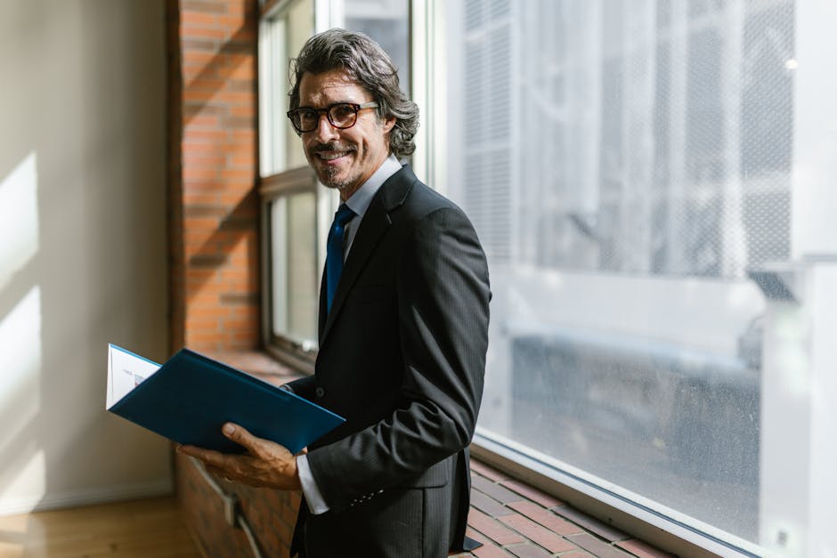 Smiling businessman in a black suit holding a folder by a window indoors.